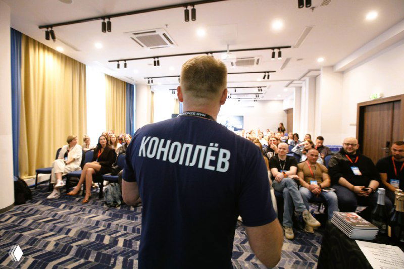 Ведущий в темной футболке с крупной надписью на спине выступает перед заполненной аудиторией в конференц-зале; участники сидят в рядах и слушают.