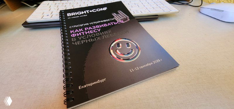 Рабочая тетрадь конференции на столе: видна черная обложка с логотипом и названием, блокнот рядом с клавиатурой и документами, наталкивает на мысли о подготовке ивента.