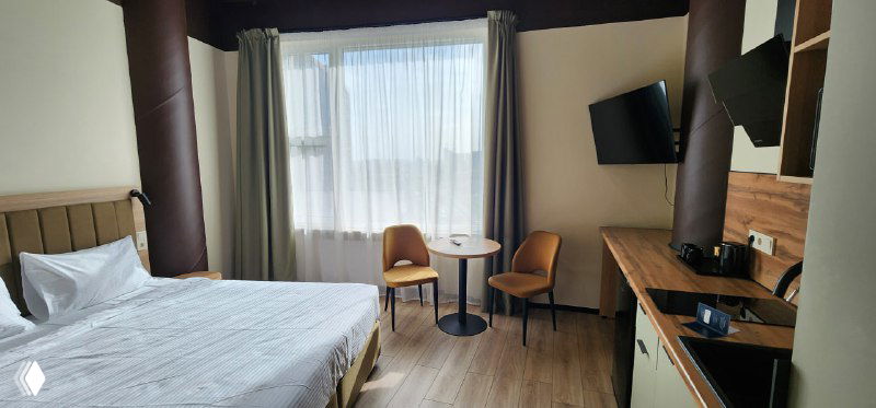 Интерьер номера отеля Idera: двуспальная кровать, столик у окна, компактная кухня с мойкой и большой телевизор на стене.