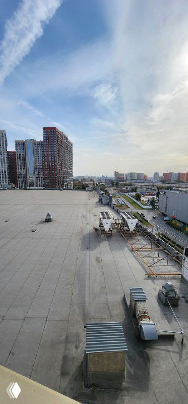 Вид из окна номера на соседние жилые кварталы и крыши, городской пейзаж с техникой на кровле и широким небом над районом.