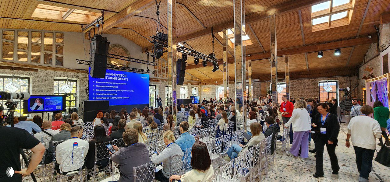 Зал с аудиторией: участники на прозрачных стульях, сцена с большим экраном и слайдами, атмосфера профессиональной конференции и обмена опытом.