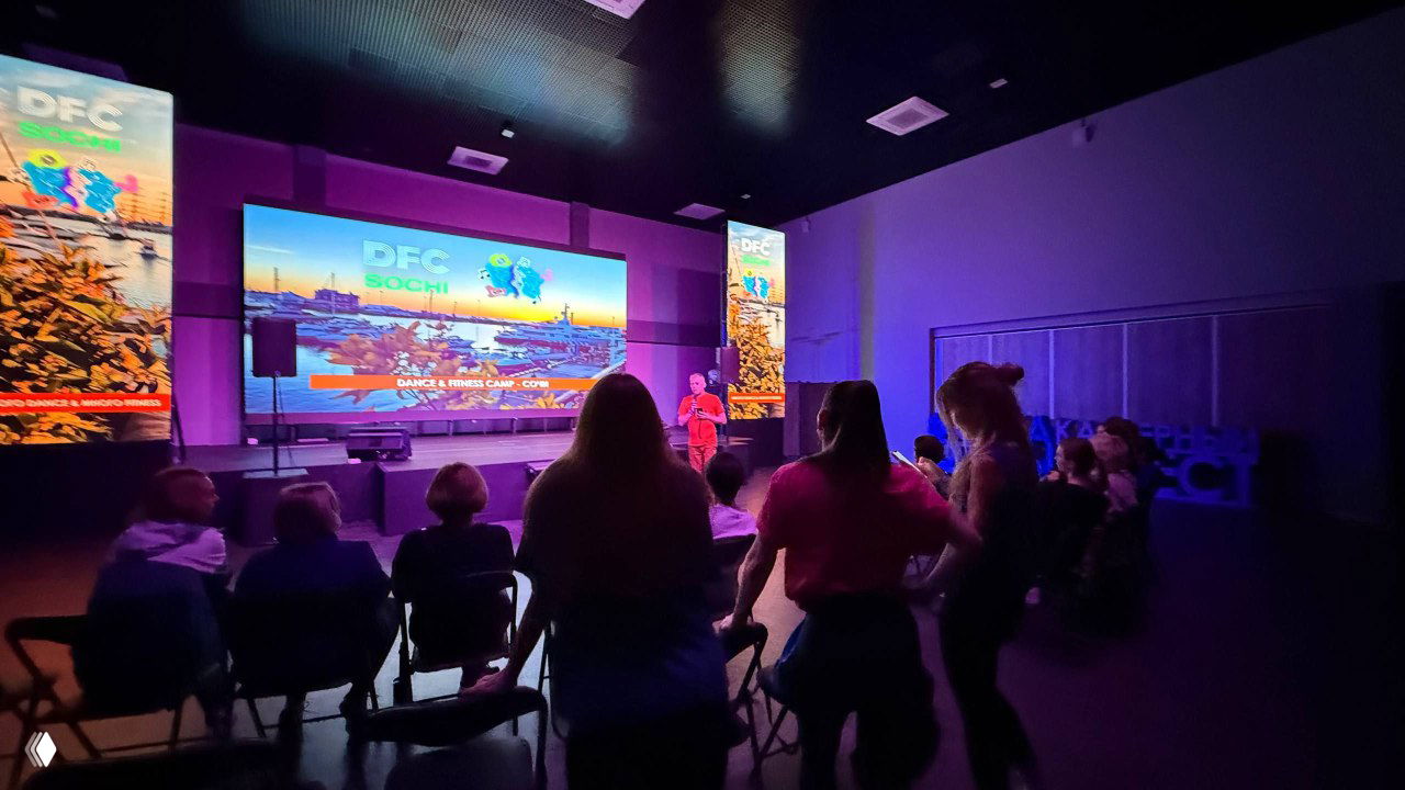 Большой зал DFC Sochi с сценой и большим экраном: ведущий выступает, логотип DFC на экране, участники сидят и стоят в полумраке аудитории.