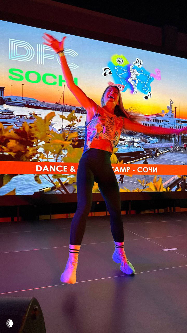 Преподаватель или участница DFC Sochi на сцене на фоне яркого экрана с надписью DFC SOCHI, динамичная поза и вечерняя подсветка.