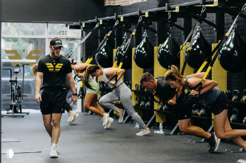 Интерьер студии Earn Your Stripes: группа людей выполняет функциональные упражнения в зале с военизированным брендингом, тренер контролирует процесс тренировки