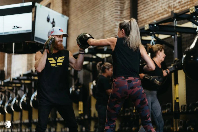 Тренер корректирует технику участницы с боевым фитнес-мячом; на заднем плане — элементы декора и логотип, создающие атмосферу «команды» и дисциплины