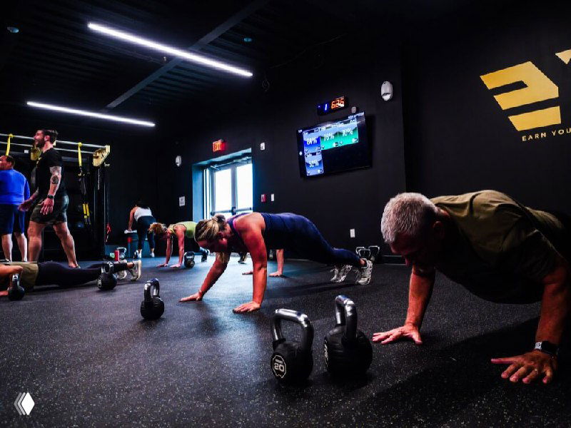 Участники выполняют упражнения с гирями и отжимания в ряд; помещение оформлено под командный функциональный класс с экраном и фирменным логотипом