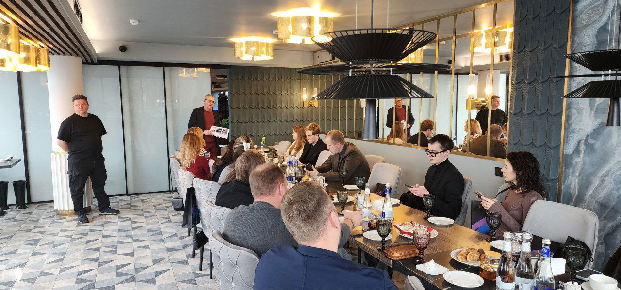 Деловая встреча участников стажировки за длинным столом: люди общаются, пьют кофе, на столе заметны бутылки воды и печатные презентации программы.