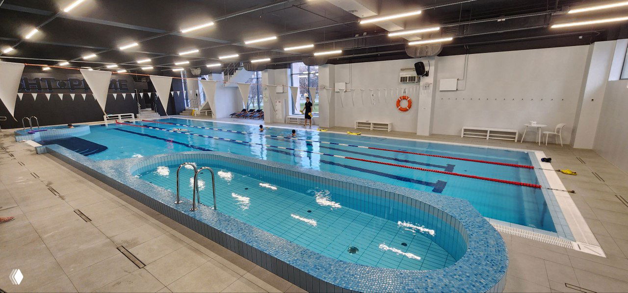 Интерьер действующего фитнес-клуба с большим бассейном и дорожками для плавания, чистая плитка, окна и просторное помещение для тренировок.