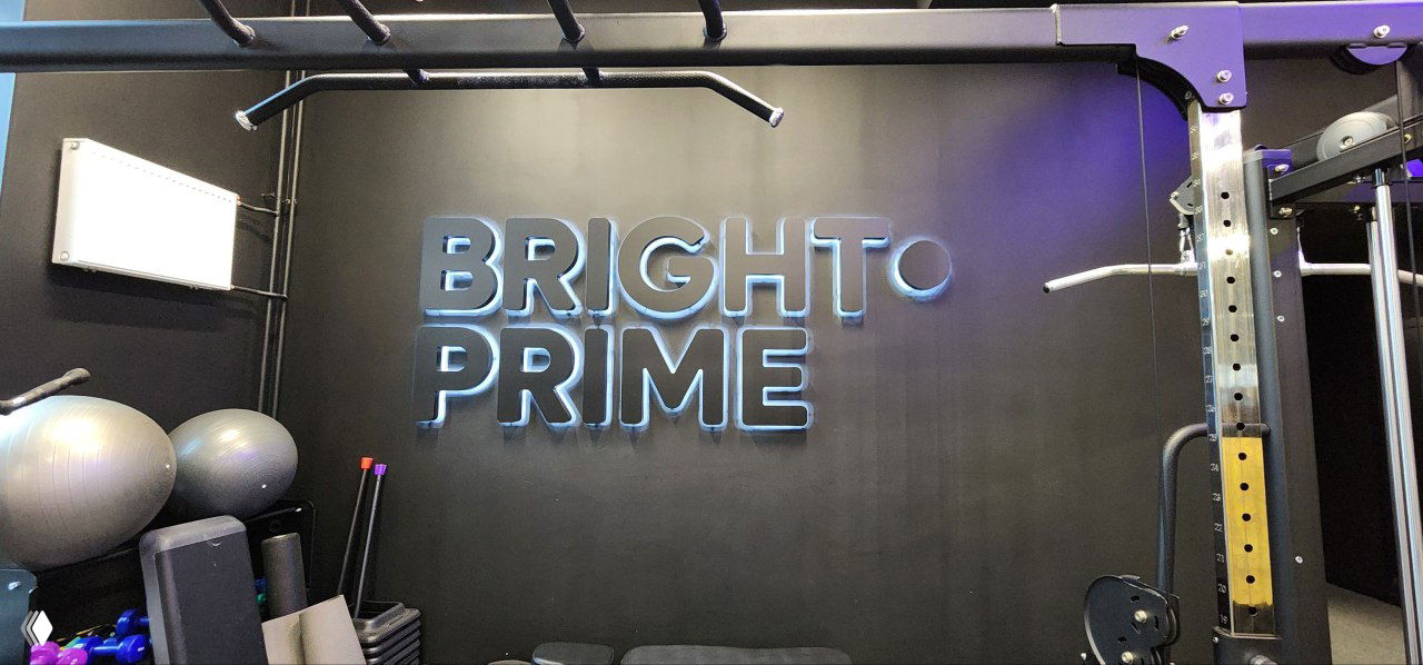 Тренажёрный уголок с яркой неоновой вывеской BRIGHT PRIME на тёмной стене, силовое оборудование и фитнес-инвентарь на переднем плане.