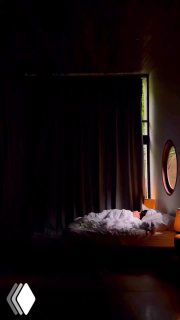 Темное фото гостиничного номера с зашторенным окном, неопрятно заправленной кроватью, круглым зеркалом и настольной лампой в углу интерьера.