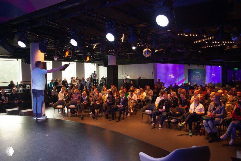 Спикер выступает на сцене перед большой заполненной аудиторией; участники сидят в креслах, в зале видно сценическое и конференц-освещение.