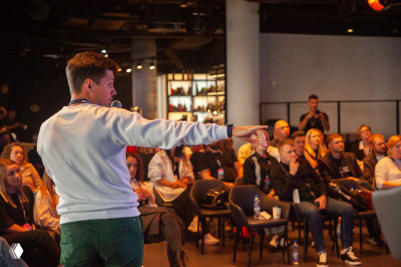 Докладчик указывает рукой на зал во время презентации; участники внимательно слушают, некоторые делают пометки, фокус на взаимодействии с аудиторией.