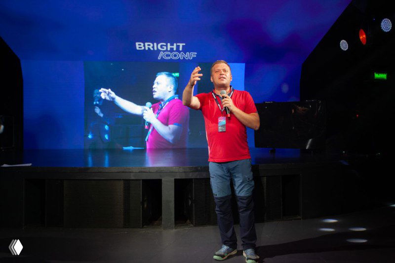 Ведущий в красной футболке на сцене Bright Conf на фоне большого экрана с трансляцией и логотипом мероприятия, дух профессиональной конференции.