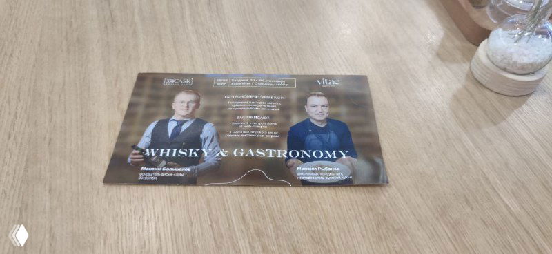 Приглашение на гастрономический бранч и дегустацию виски, расположенное на деревянном столе; видна карточка с текстом и логотипами, часть интерьера клуба.