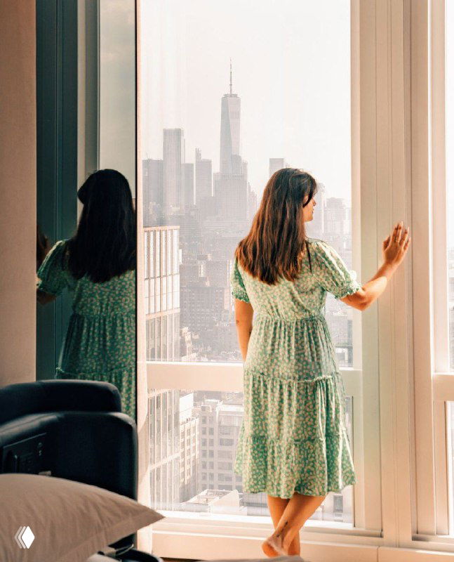 Гостья в лёгком платье у окна номера, смотрящая на городской пейзаж — кадр, передающий личные впечатления и атмосферу.
