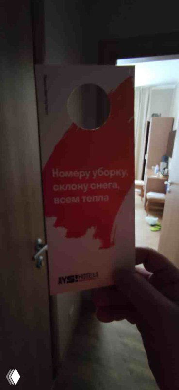 Держатель на дверь номера Ays с красно‑белой графикой и надписью о уборке; на фоне видно внутреннюю часть номера и часть коридора отеля.