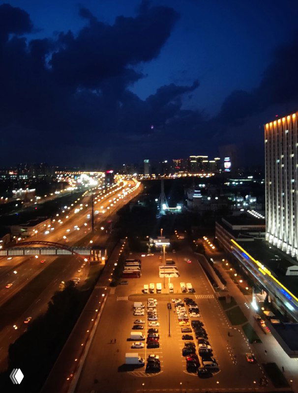 Ночной вид из окна 20 этажа отеля: освещённая автостоянка и шоссе, светящиеся здания Москвы, ярко подсвеченный фасад отеля справа на кадре.