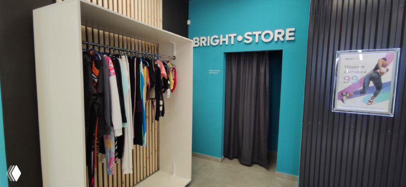 Примерочная и витрина фирменного мерча в новом клубе Bright Fit: стойка с одеждой, вход в примерочную и рекламный плакат на стене.