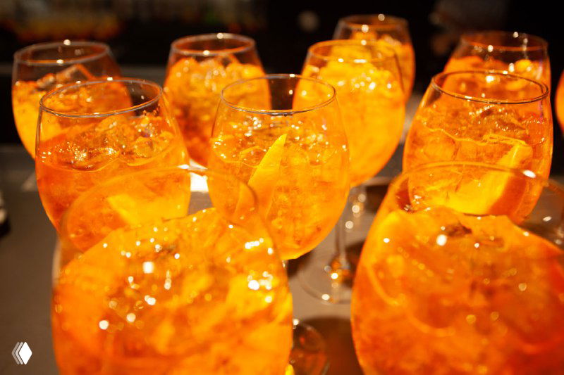 Бокалы с апероль-коктейлем на фуршете конференции: яркие оранжевые напитки, лед и дольки апельсина в официозной атмосфере ивента.