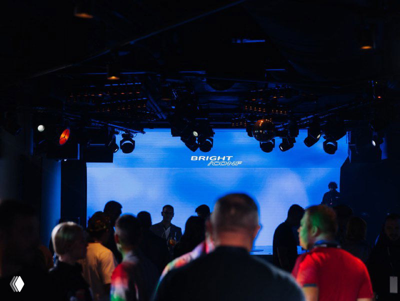 Зрительный зал ночного клуба перед началом деловой программы: сцена, проекционный экран и собравшаяся аудитория в полумраке.