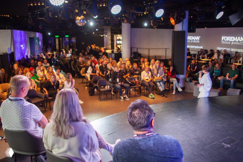 Панорамный кадр с выступающей сценой и полной зрительской залой — деловая сессия в формате конференции в клубе.