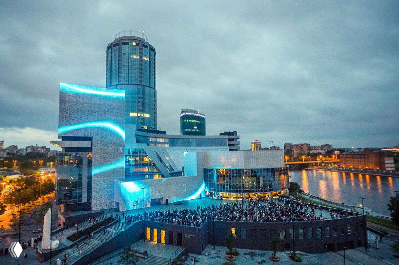 Вид на набережную города вечером с подсвеченными зданиями и собравшейся аудиторией на открытом пространстве — городской фон мероприятия.