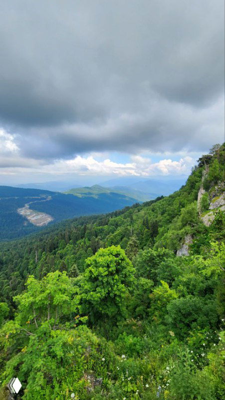 Вертикальный кадр горного склона и зелёной растительности в Адыгее: вид с утёса на лесистые хребты, долины и тяжелое облачное небо.