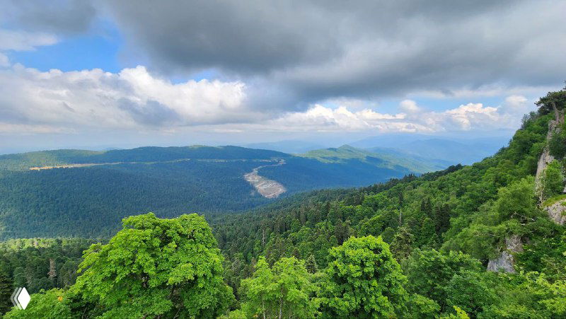 Широкий пейзаж горной Адыгеи с лесными массивами и дальними хребтами: видовые площадки, глубокие впадины и переменчивая погода над ландшафтом.