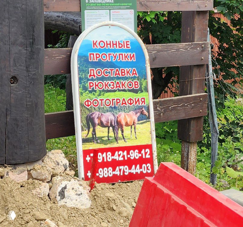 Рекламный штендер с надписями «Конные прогулки, доставка рюкзаков, фотография» у деревянного забора на плато Лаго-Наки.