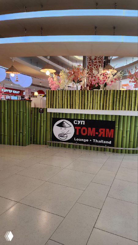 Вывеска фудкорта с надписью «Суп Том Ям» в торговом центре; точка с бамбуковым забором, декоративными растениями и пустой зоной перед ней.