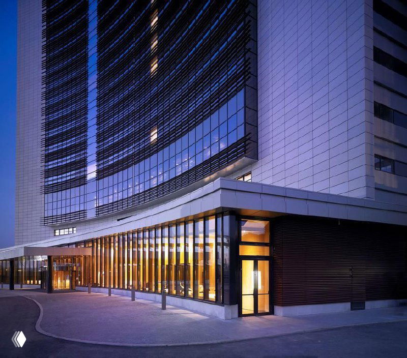 Угловой фасад большого городского отеля с ночным освещением и стеклянными поверхностями на фоне синего неба — гостиница как место размещения участников конференции