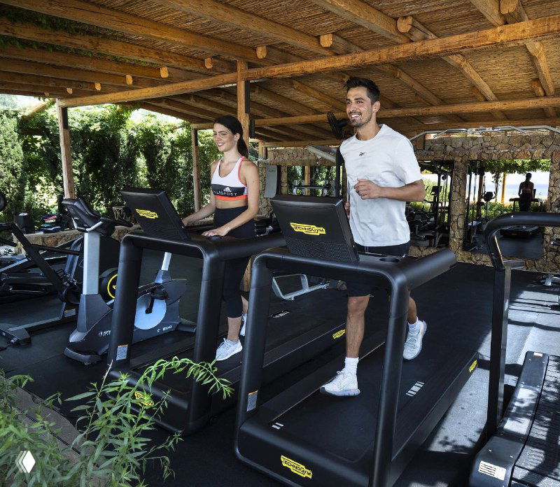Открытый тренажёрный уголок на территории отеля под навесом: беговые дорожки и зелёные насаждения вокруг, два человека занимаются кардио.