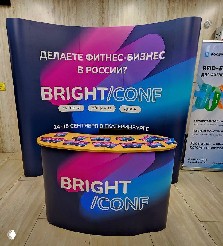 Рекламный стенд Bright Conf с ярким баннером «Делаете фитнес-бизнес в России?» и столом с раздаточными материалами для привлечения участников конференции.