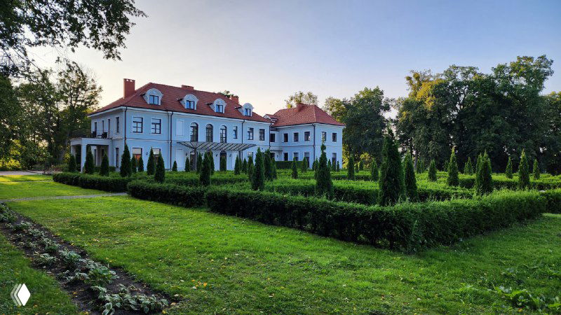 Фасад загородного отеля-ресторана «Усадьба», ухоженный парк перед зданием и аккуратные зелёные стрижки на фоне вечернего света.