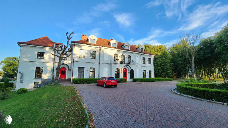 Вид на второй корпус отеля «Усадьба», мощёная подъездная зона, красный автомобиль у входа и ясное голубое небо.