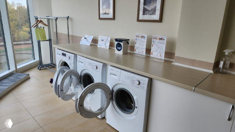 Интерьер гостевой прачечной отеля 4 Elements (Киров): три фронтальные стиральные машины, сушилка, столешница и аккуратная зона у окна.
