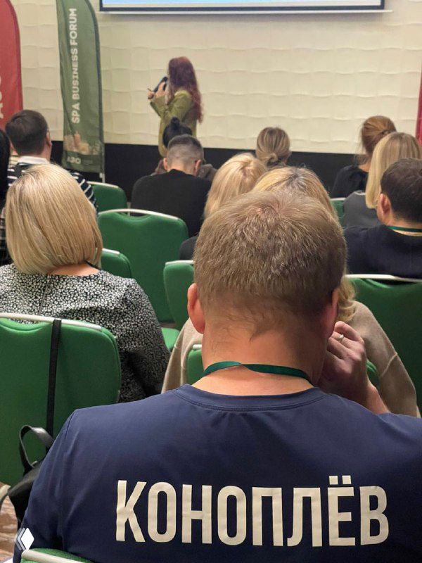 Конференц‑зал спа‑форума: спикер у экрана, аудитория в зелёных креслах, на переднем плане участник в футболке «Коноплёв».