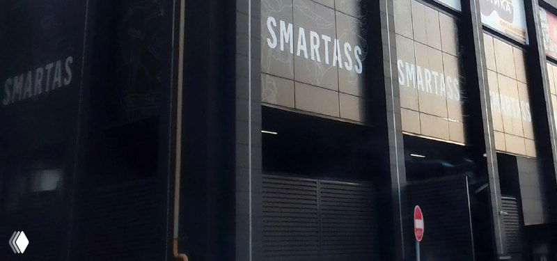 Чёрный фасад здания с крупной вывеской «SMARTASS», городской вид у входа фитнес-клуба, припаркованные машины, молодой стиль.