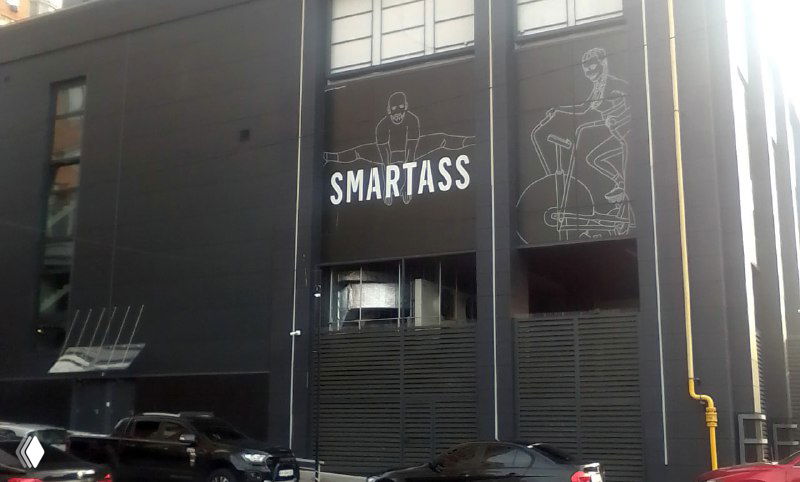 Фасад и вывеска «SMARTASS» на чёрном здании фитнес-клуба; контурные графические рисунки на стене, улица и припаркованные автомобили.