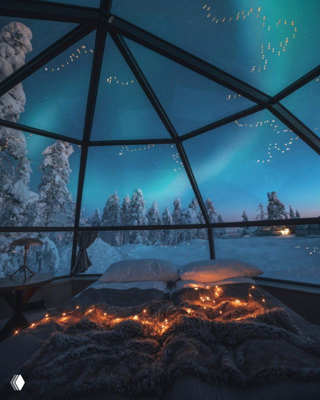 Вид из стеклянного купола на кровать с тёплыми пледами, заснеженный лес и северное сияние — тишина и уединение зимой.
