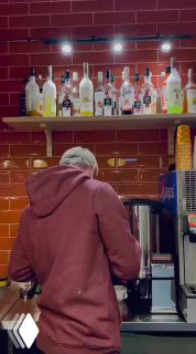 Человек в худи спиной за кофейной станцией в кафе; на фоне полки с бутылками, красная плитка и кофейное оборудование.