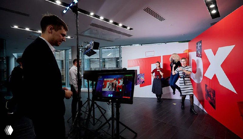 Интерьерная фотозона с красной брендированной стеной и большой буквой TRIX, сотрудники и гости взаимодействуют с экраном и техникой для съёмки.