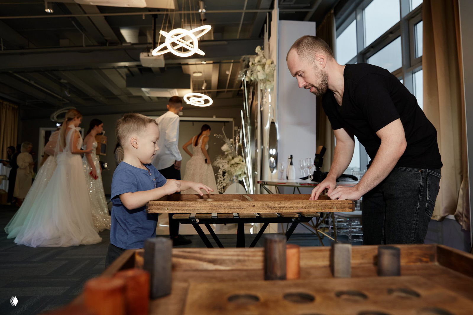 Игроки на фоне свадебного мероприятия: мальчик и взрослый играют за столом с деревянными игровыми элементами, атмосфера праздника.