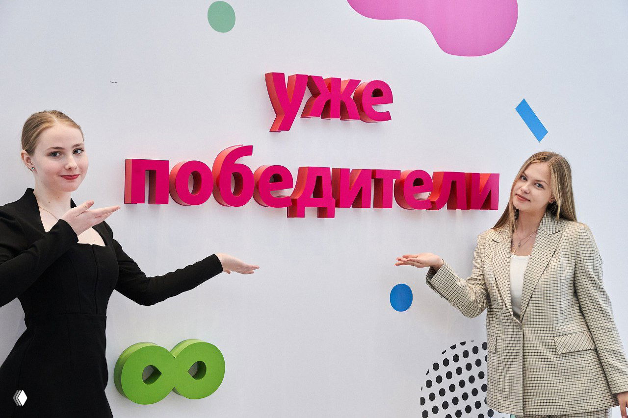 Две участницы показывают на яркую надпись «уже победители» на тематическом стенде; фотография передает атмосферу торжественной церемонии и оформления пространства.