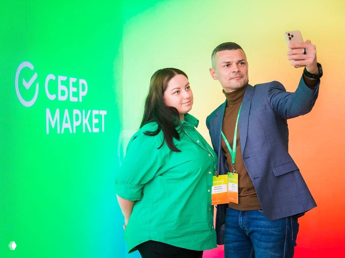 Двое гостей делают селфи у яркой LED‑фотозоны с логотипом «Сбер Маркет»: цветной градиентный фон мероприятия.