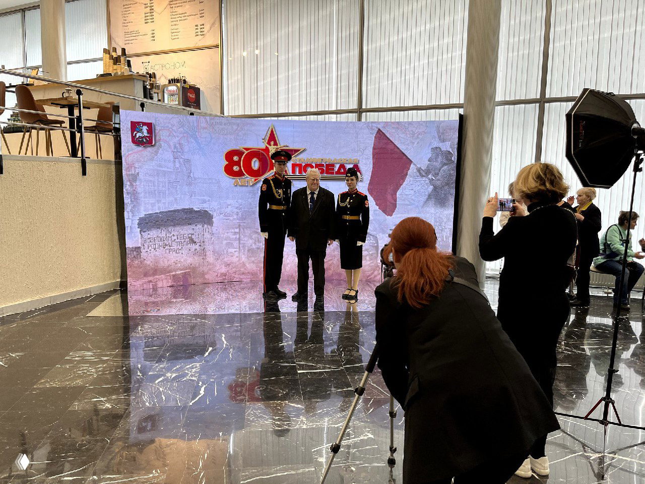 Фотограф снимает ветерана и почётный караул перед LED-фотозоной с символикой «80 Победа», видна фототехника и отражения на мраморном полу