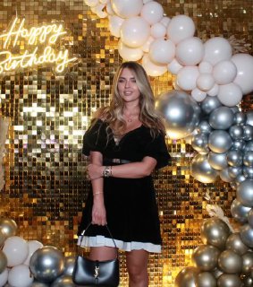 Женщина рядом с золотой блестящей фотозоной, украшенной белыми и серебряными шарами и неоновым знаком, пример оформления для мероприятия и фотосессии.