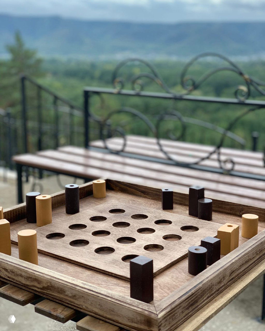 Деревянная крафтовая настольная игра с круглыми лунками и цилиндрическими фишками на столе, с размытым фоном пейзажа в кадре.