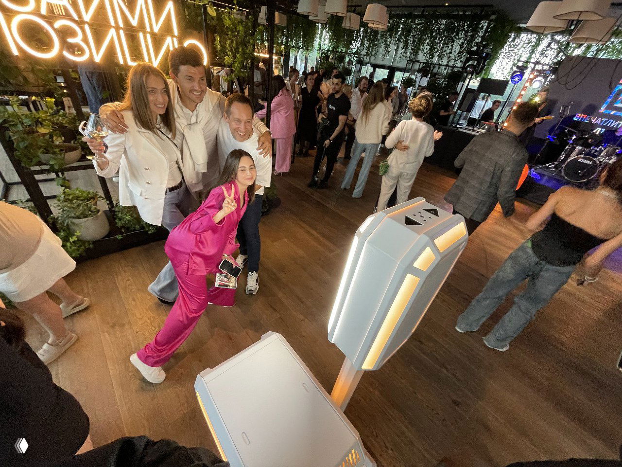 Группа гостей на мероприятии делает фото рядом с современной белой фотостойкой MixUp в оформленном пространстве с неоновыми вывесками и растениями.