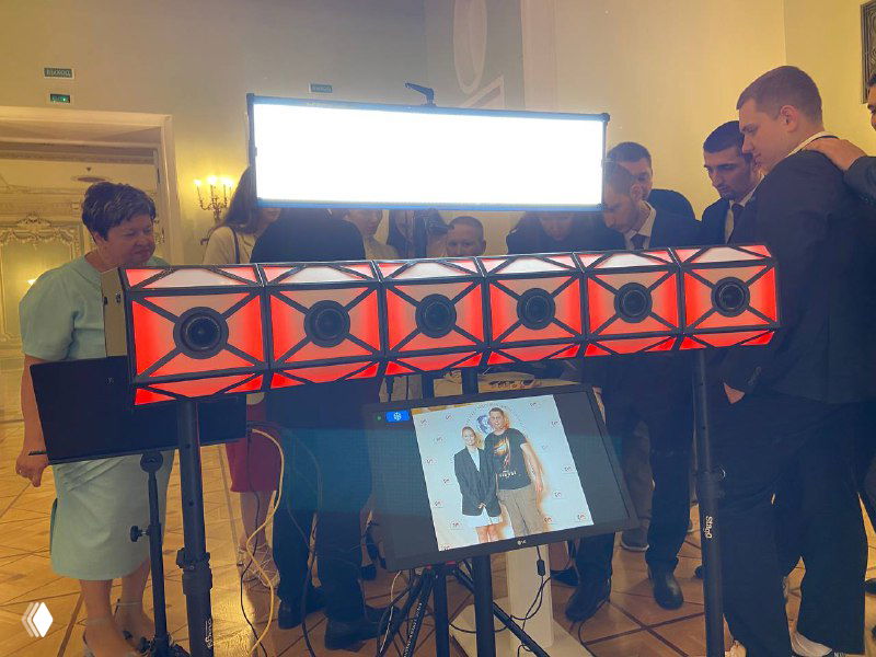 Компактная 3D‑фотостойка с яркой подсветкой и экраном, вокруг — гости мероприятия, видно оборудование и место установки в зале